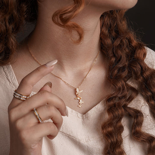 A close-up shot of a person's chest and neck shows a delicate gold chain necklace with a cluster of three small butterfly-shaped pendants, each sparkling with clear gemstones. The person is also wearing several gold rings on their finger.