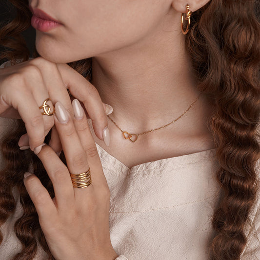 A close-up shot of a person wearing a beige top. A delicate gold chain necklace is visible around their neck, featuring two small, interlocking heart pendants. The person also wears gold rings and hoop earrings.