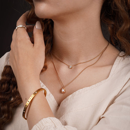 A close-up shot of a person's chest, neck, and hand. The person is wearing a beige top. A delicate, layered necklace set is visible, with a small, single-stone charm on the upper chain and a charm with a teardrop-shaped white pearl on the lower chain. A gold bangle with Roman numerals is on their wrist, and a bold ring with pearls is on their finger.