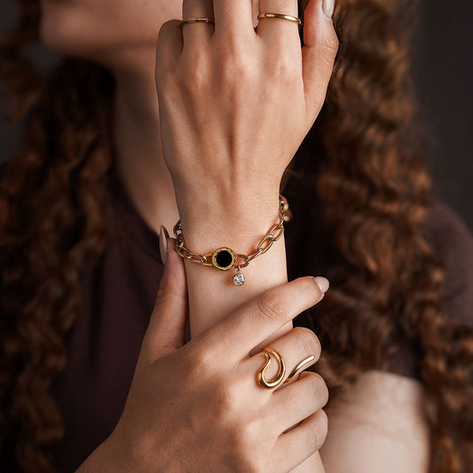 A close-up shot of a person's hands and arms, showcasing a variety of gold-toned jewelry. On the wrist is a chunky chain bracelet with a black and gold circular charm from which a small gemstone hangs. The fingers are adorned with several rings: a simple thin band, a thicker polished band, and a bold, wave-like ring.