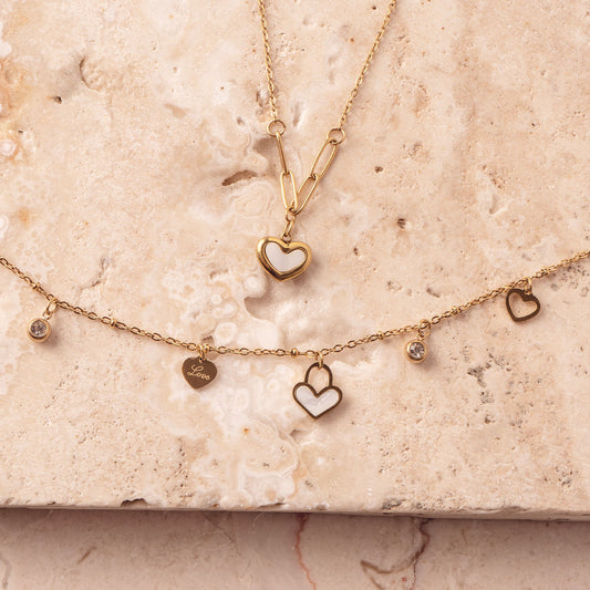 Two delicate 18k gold plated necklaces arranged on a light-colored marble surface. The top necklace features a Violet Petal Pendant, while the bottom necklace showcases multiple Amara Love Charms, including heart-shaped and sparkling designs.