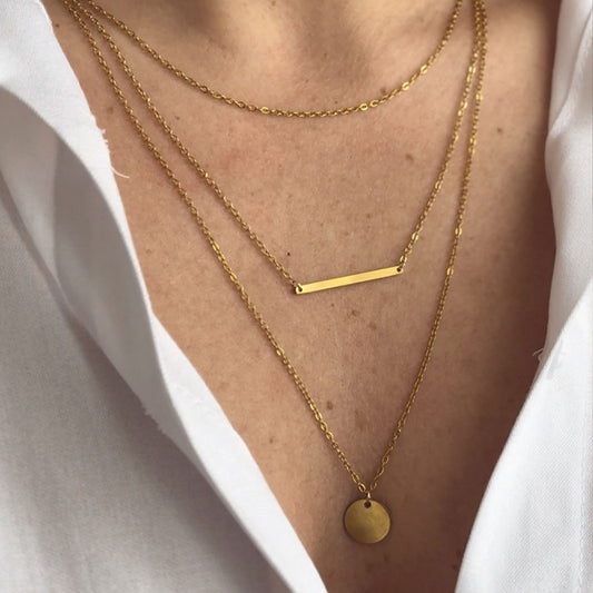 A close-up of a person's chest, showcasing three layered 18k gold plated Camellia Classic Chain necklaces. One features a sleek horizontal bar pendant, another a simple circular disc pendant, and the top chain is unadorned, all resting elegantly against a white collared shirt.