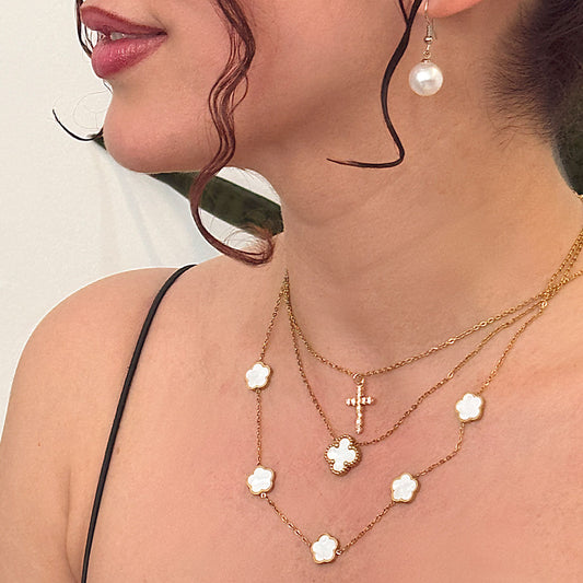 A close-up of a person's neckline adorned with a beautiful layered arrangement of 18k gold plated necklaces. This includes a Luminous Faith Pendant in the shape of a sparkling cross, a Celestia Bloom Necklace with multiple white floral charms, and a Sophia Timeless Clover Necklace, completing a sophisticated look.