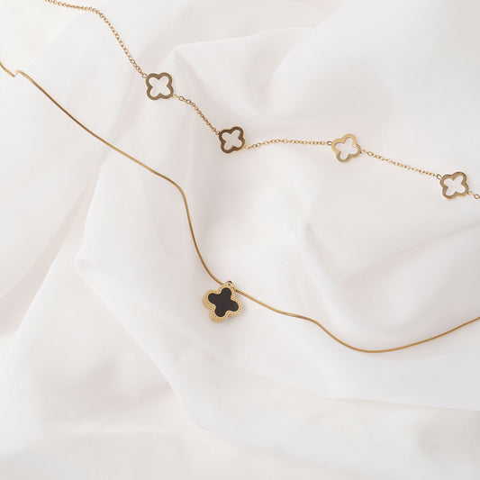 Two elegant 18k gold plated necklaces displayed on a soft, draped white fabric. The lower necklace features a single Serena Clover Pendant with a dark center, while the upper necklace showcases multiple open clover charms.