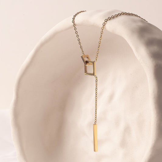 An elegant 18k gold plated Jasmine Grace Necklace, featuring a unique lariat design with an open square link and a sleek rectangular bar pendant, gracefully draped over a textured white sculpture.