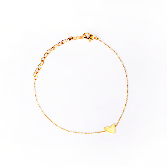 The Anna Heart Bracelet, a delicate gold-toned chain, is arranged in a circle on a clean white background. A small, polished, flat heart charm is positioned along the chain, giving the bracelet a sweet and minimalist design.