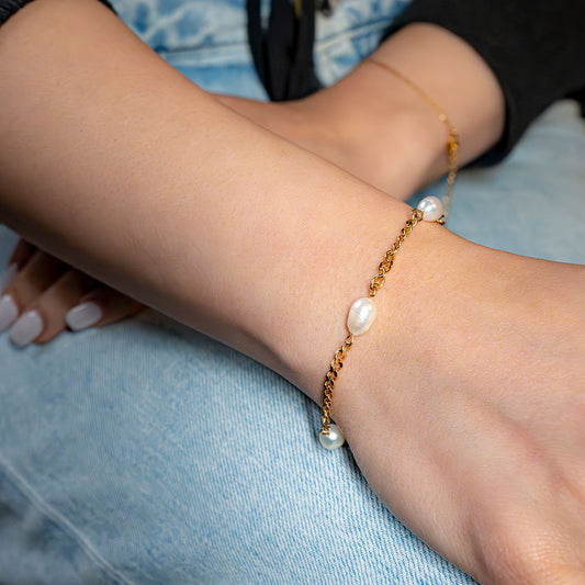 A close-up view captures a person's arms, with one wrist adorned by the Aura Pearl Bracelet. This elegant gold-toned bracelet features a delicate chain beautifully interspersed with several lustrous, irregularly shaped white pearls, resting gently against a backdrop of light blue denim.