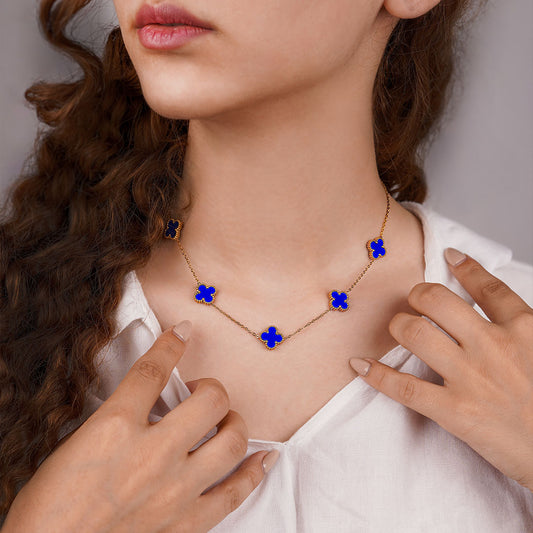 A person wearing a white shirt is shown from the chest up, wearing the Aurora Clover Pendant. The gold necklace features five striking blue clover pendants spaced along the delicate chain.