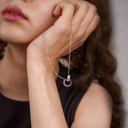 A close-up shot of a person's hand holding the Aurora Trend Necklace. The silver-toned chain leads to a centerpiece of two interlocking rings; one has Roman numeral engravings, and the other is encrusted with small, clear gemstones.