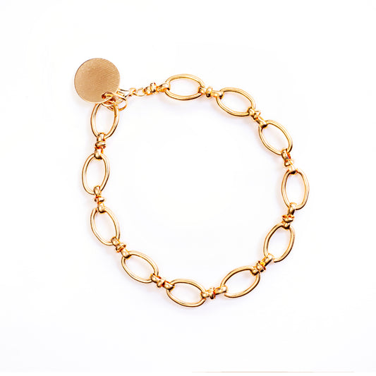 The Azara Oval Chain Bracelet is presented against a bright white background, forming a large oval shape. This gold-toned bracelet is comprised of numerous interconnected, polished oval links, and includes a small, circular disc charm near its clasp.