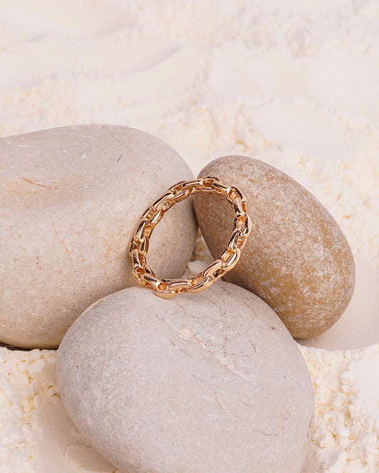 The Elara Chain Ring is showcased resting on a smooth white stone, surrounded by other stones and a bed of fine white powder. The gold-toned ring has a delicate, continuous chain of interconnected oval links.