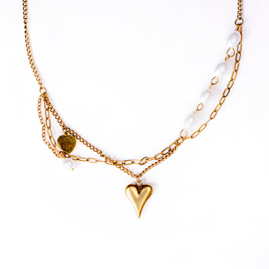 A studio shot on a white background displays the Elara Heirloom Necklace. This multi-layered gold-toned necklace features various chains, including one with interspersed white pearls, and is adorned with charms such as a small gold coin, a dangling pearl, and a central, puffed gold heart pendant.