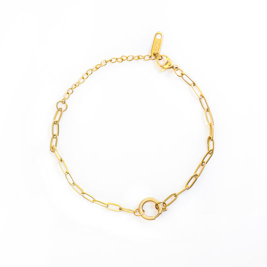 The Elara Loop Bracelet is laid out in a circular shape on a white background. This gold-toned bracelet features an elongated paperclip-style chain that connects to a prominent, polished circular clasp at its center, with an extender chain near the lobster claw closure.