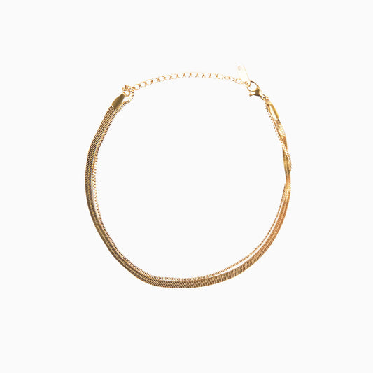 A studio shot on a white background displays the Flora Herringbone Bracelet. This gold-toned bracelet features a sleek, flat, and flexible herringbone chain, arranged in a circular shape with an extender chain and clasp visible at the top.