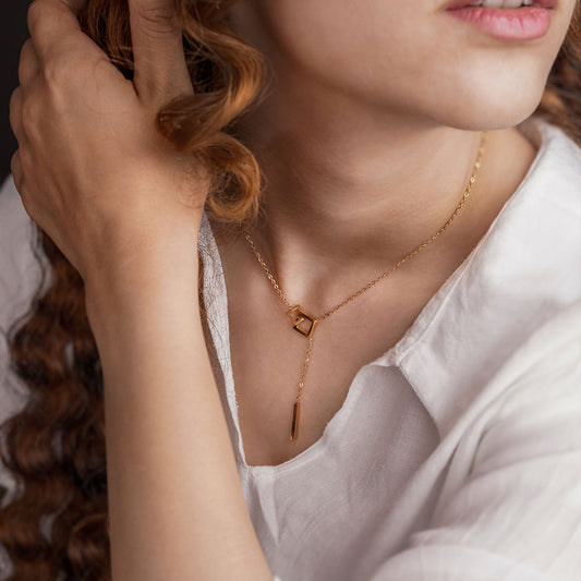 A woman wearing a white collared shirt and the Gold Plated Jasmine Grace Necklace, which features a lariat and bar drop pendant, with one hand in her curly hair.