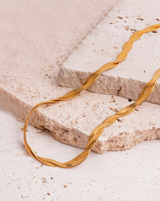 The Lumina Herringbone Twist Necklace is showcased on a beige, textured stone surface. This gold-toned necklace features two intertwined herringbone chains, their brilliant links twisting together to create a striking textural contrast against the rough rock.