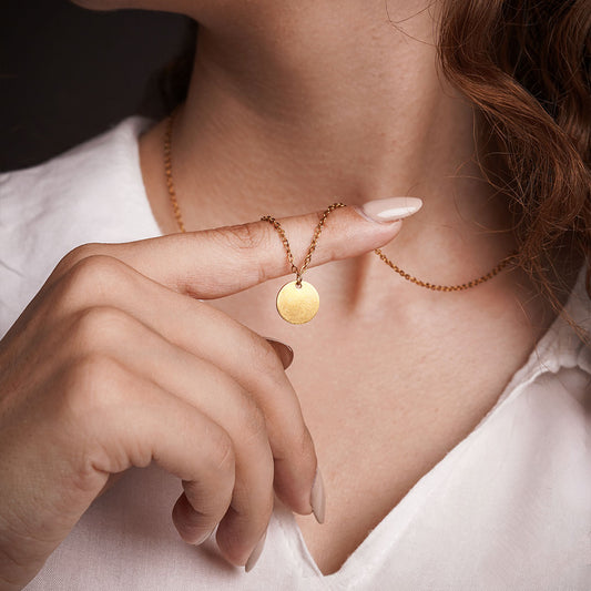 A close-up shot focuses on a person's hand gently holding the Luna Disc Necklace. The delicate gold chain and its simple, gleaming disc pendant are showcased against a white shirt, with the necklace resting just below the collarbone.
