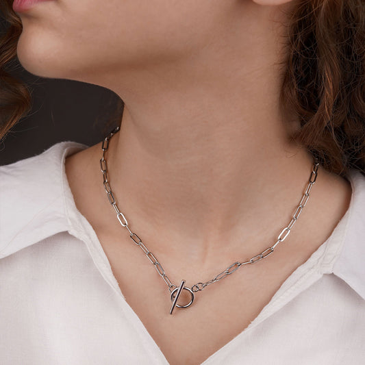 close-up side profile of a person wearing a white collared shirt. The Lustrous Arc Necklace, a silver-toned necklace with a paperclip chain and a prominent toggle clasp, is worn just below the collarbone.