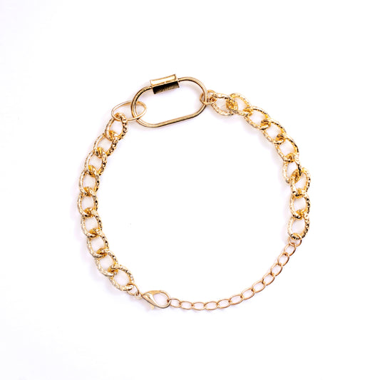 The Rhea Link Bracelet is showcased forming a circle on a white background. This gold-toned bracelet features a chunky, textured chain on one side connected by a large, smooth oval carabiner-style clasp, leading to a finer chain on the other side.