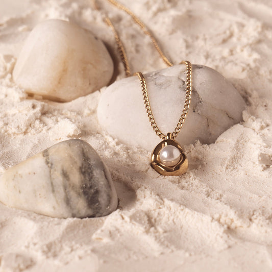 The Saffron Luxe Necklace by Delta Jewel, crafted from high-quality stainless steel with a luxurious gold plating, featuring a sculptural pendant holding a luminous pearl, styled on soft sand among smooth white stones.