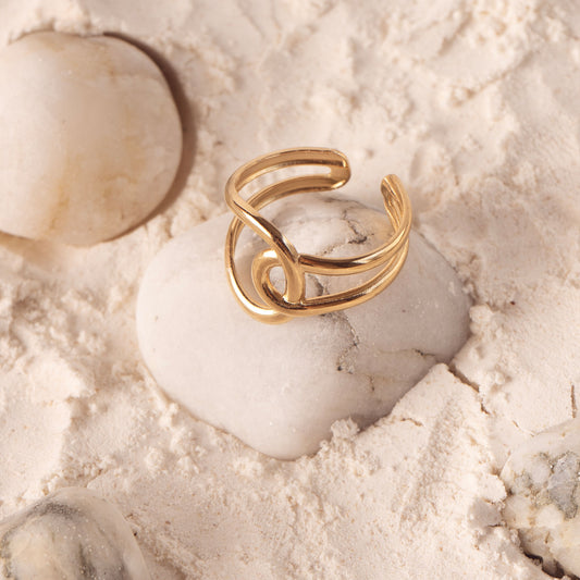 Seraphine Charm Ring by Delta Jewel, a sculptural gold-plated ring with a fluid knot design, displayed on white sand and smooth stones.