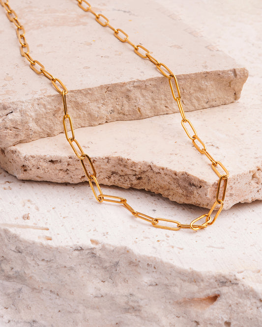 The Serena Paperclip Necklace is showcased on a beige, textured stone surface. The gold-toned necklace features a chain of sleek, elongated links that are draped across the rock.