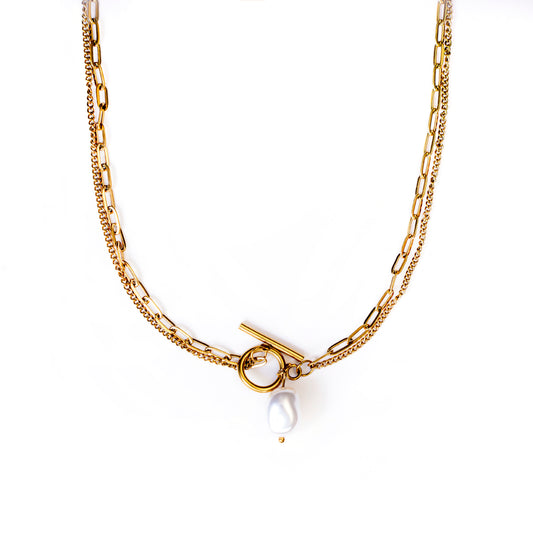 A studio shot on a clean white background displays the Solara Toggle Pearl Necklace. This gold-toned, layered necklace features two distinct chains (one with elongated links, one finer) that meet at a prominent toggle clasp. A large, irregularly shaped white baroque pearl dangles from the toggle clasp.
