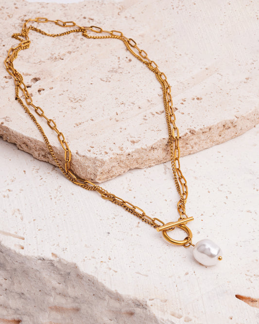 The Solara Toggle Pearl Necklace is elegantly draped over a textured, beige stone surface. The layered gold necklace features two distinct chains that meet at a prominent toggle clasp, which holds a single, lustrous baroque pearl.