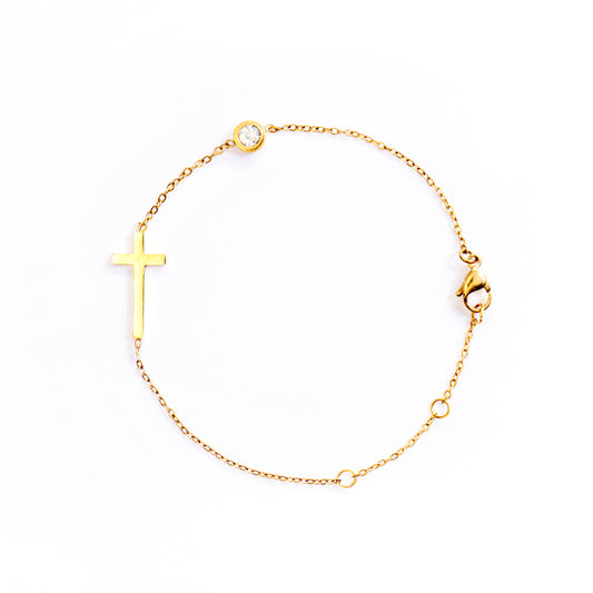 The Stella Grace Bracelet is presented in a studio shot on a white background, forming a circle. This gold-toned bracelet features a minimalist, flat cross charm positioned along the chain, with a small, clear round gemstone set further along the chain.