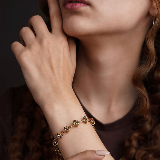 The Valerie Knotted Bracelet is presented in a studio shot on a clean white background, arranged in a circular shape. The gold-toned bracelet is composed of a series of elongated links that are intertwined with smaller knotted links, with a clasp and extender chain visible at the top.