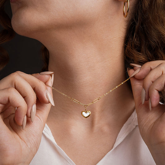A close-up shot of a person wearing the Violet Petal Pendant, a gold necklace with a white-inlaid heart pendant. The person's hands are holding the necklace's mixed-link chain, and small gold hoop earrings are visible on their ears.