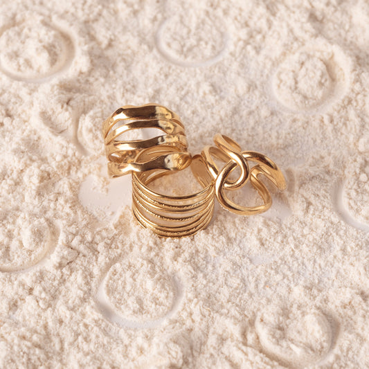 3 rings by Delta Jewel which are Amelia Radiant Ring and Mila Zircon Ring, all with a bold gold-plated band with parallel grooves and a polished finish, displayed on textured sand alongside other statement rings.