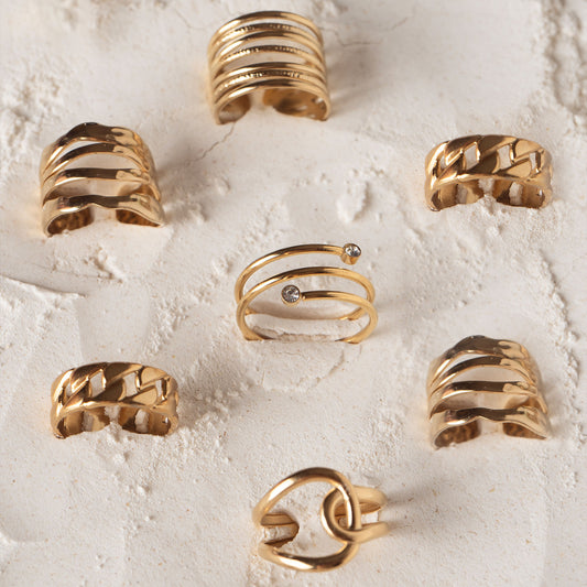 A set of rings including Amelia Radiant Ring, Delta Clover Ring and Mila Zircon Ring by Delta Jewel, a sleek gold-plated spiral ring accented with two clear stones, displayed at the center of a curated flat lay of gold-plated rings on textured sand.