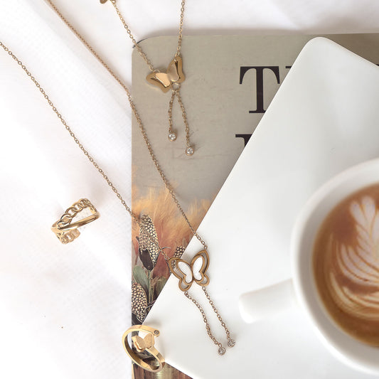 A flat lay display featuring an 18k gold plated Juliette French Ring and an 18k gold plated Willow Butterfly Charm necklace, elegantly arranged with a cup of coffee and a book.