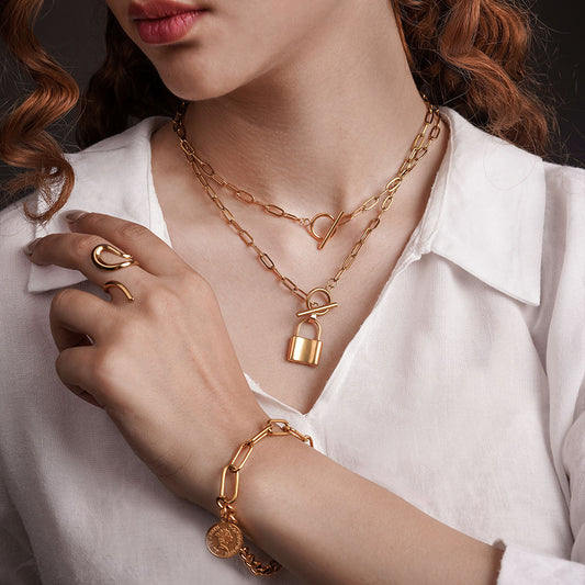 A person wearing a white collared shirt displays several pieces of gold-toned jewelry. Around their neck is a layered necklace set: the upper chain has a toggle clasp closure, while the lower chain also features a toggle clasp from which a small, polished lock pendant hangs. On their hand, a bold, sculptural ring is visible, along with a thick chain bracelet adorned with a circular coin charm.