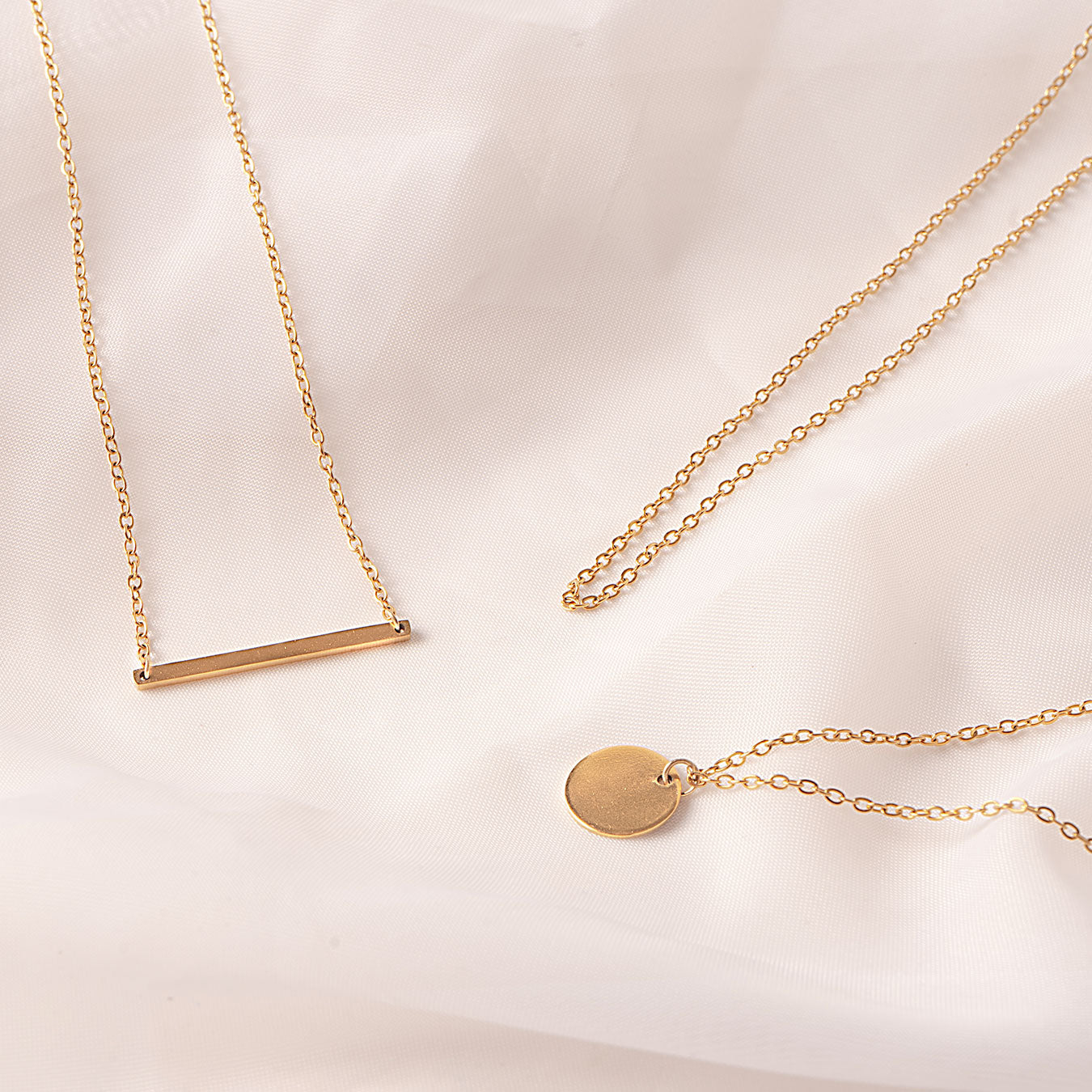 Three delicate 18k gold plated Camellia Classic Chain necklaces are artfully arranged on a soft, draped white fabric. One necklace features a sleek horizontal bar pendant, another a simple circular disc pendant, and a third chain is shown without a pendant, emphasizing the versatility of the classic chain.