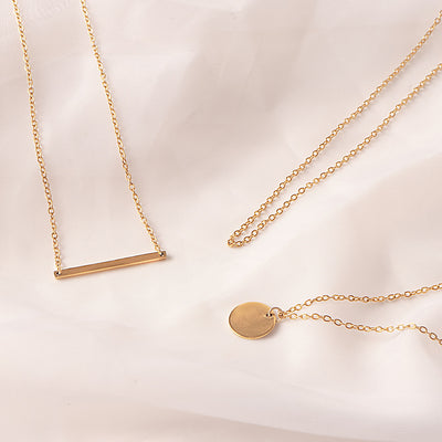 Three delicate 18k gold plated Camellia Classic Chain necklaces are artfully arranged on a soft, draped white fabric. One necklace features a sleek horizontal bar pendant, another a simple circular disc pendant, and a third chain is shown without a pendant, emphasizing the versatility of the classic chain.