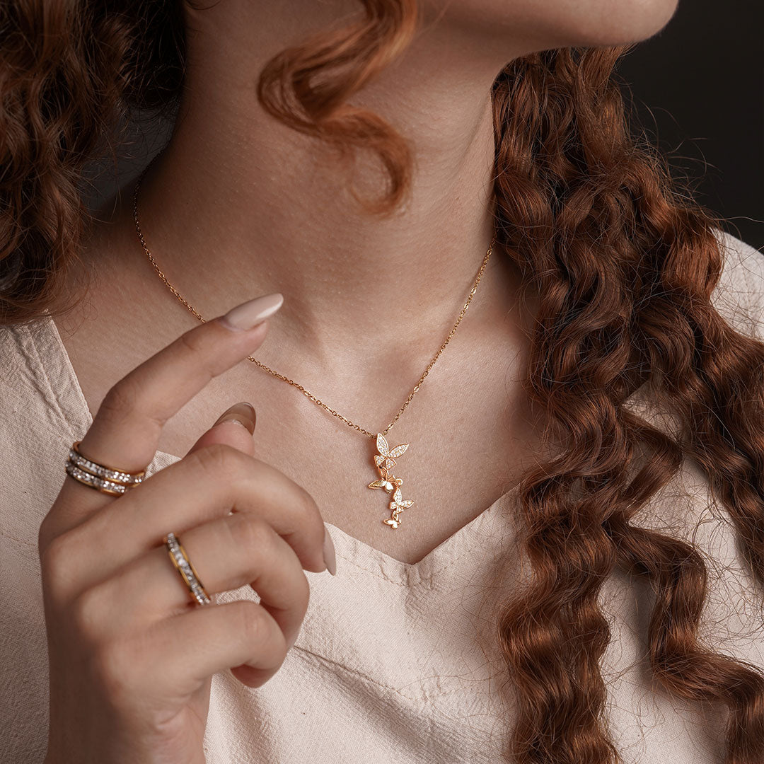 A close-up shot of a person's chest and neck shows a delicate gold chain necklace with a cluster of three small butterfly-shaped pendants, each sparkling with clear gemstones. The person is also wearing several gold rings on their finger.