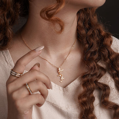A close-up shot of a person's chest and neck shows a delicate gold chain necklace with a cluster of three small butterfly-shaped pendants, each sparkling with clear gemstones. The person is also wearing several gold rings on their finger.