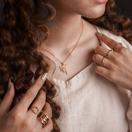 A close-up shot of a person's chest and neck, wearing a white shirt. A delicate gold chain necklace with a small, sparkling palm tree pendant is visible. The person is also wearing several gold rings on their fingers.