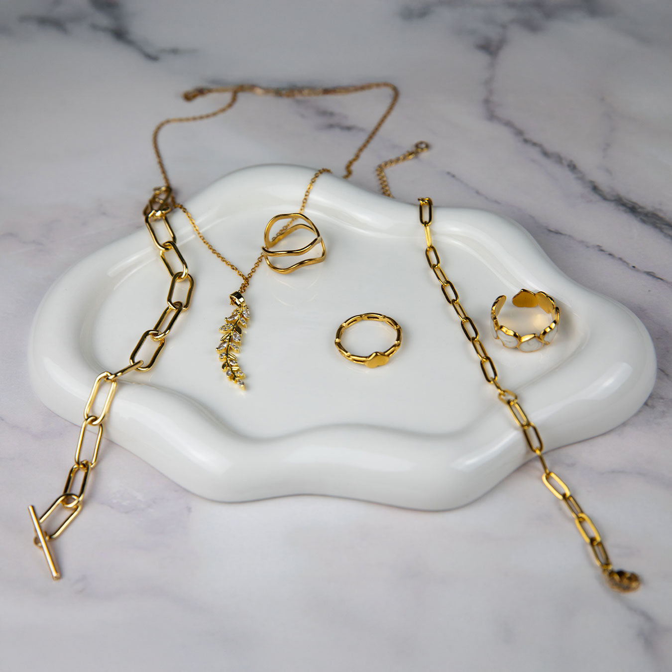 An elegant collection of 18k gold plated jewelry artfully arranged on a white cloud-shaped tray set against a marble background. The display includes a Dahlia Bloom Necklace with a delicate leaf pendant, an Aurora Grace Bracelet, a Flora Petite Band, a Dahlia Charm Ring, and an Amara Heart Ring, showcasing a luxurious golden assortment.