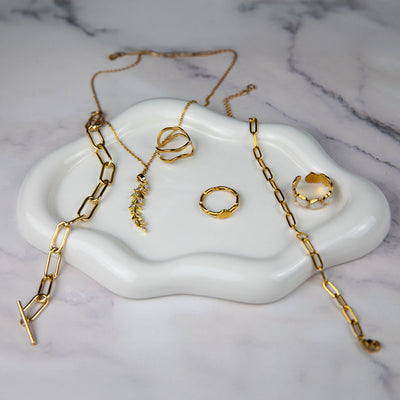 An elegant collection of 18k gold plated jewelry artfully arranged on a white cloud-shaped tray set against a marble background. The display includes a Dahlia Bloom Necklace with a delicate leaf pendant, an Aurora Grace Bracelet, a Flora Petite Band, a Dahlia Charm Ring, and an Amara Heart Ring, showcasing a luxurious golden assortment.