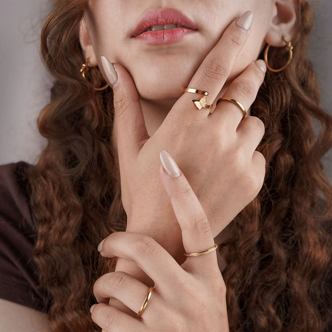 A close-up of a person's hands and face, focusing on their gold jewelry. The person wears a gold butterfly-shaped ring with a small gemstone, a simple polished gold band, and a textured band ring, as well as gold hoop earrings.