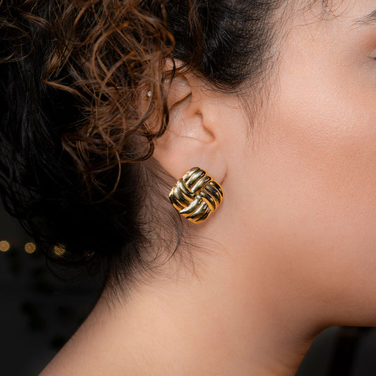 Close-up of a person's ear wearing a large, square-shaped gold stud earring with an intricate woven or knot-like design. The earring has a shiny, metallic finish.