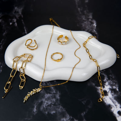 An exquisite collection of 18k gold plated jewelry elegantly arranged on a white cloud-shaped tray, set against a dark marble background. The display features a Dahlia Bloom Necklace with a delicate leaf pendant, an Aurora Grace Bracelet, a Flora Petite Band, a Dahlia Charm Ring, and an Amara Heart Ring, showcasing their radiant golden finish.