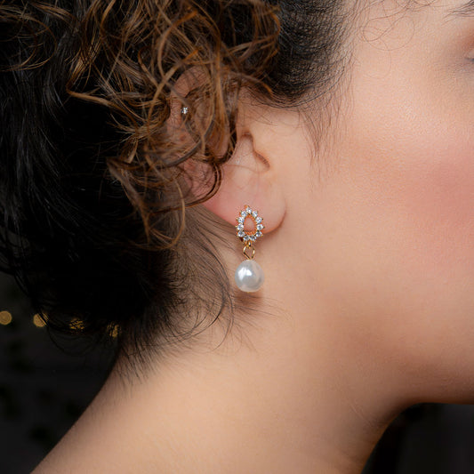 Close-up of a person's ear wearing a gold dangle earring. The earring features a top part with a pear-shaped outline adorned with small clear stones, and a single lustrous white pearl suspended from it.