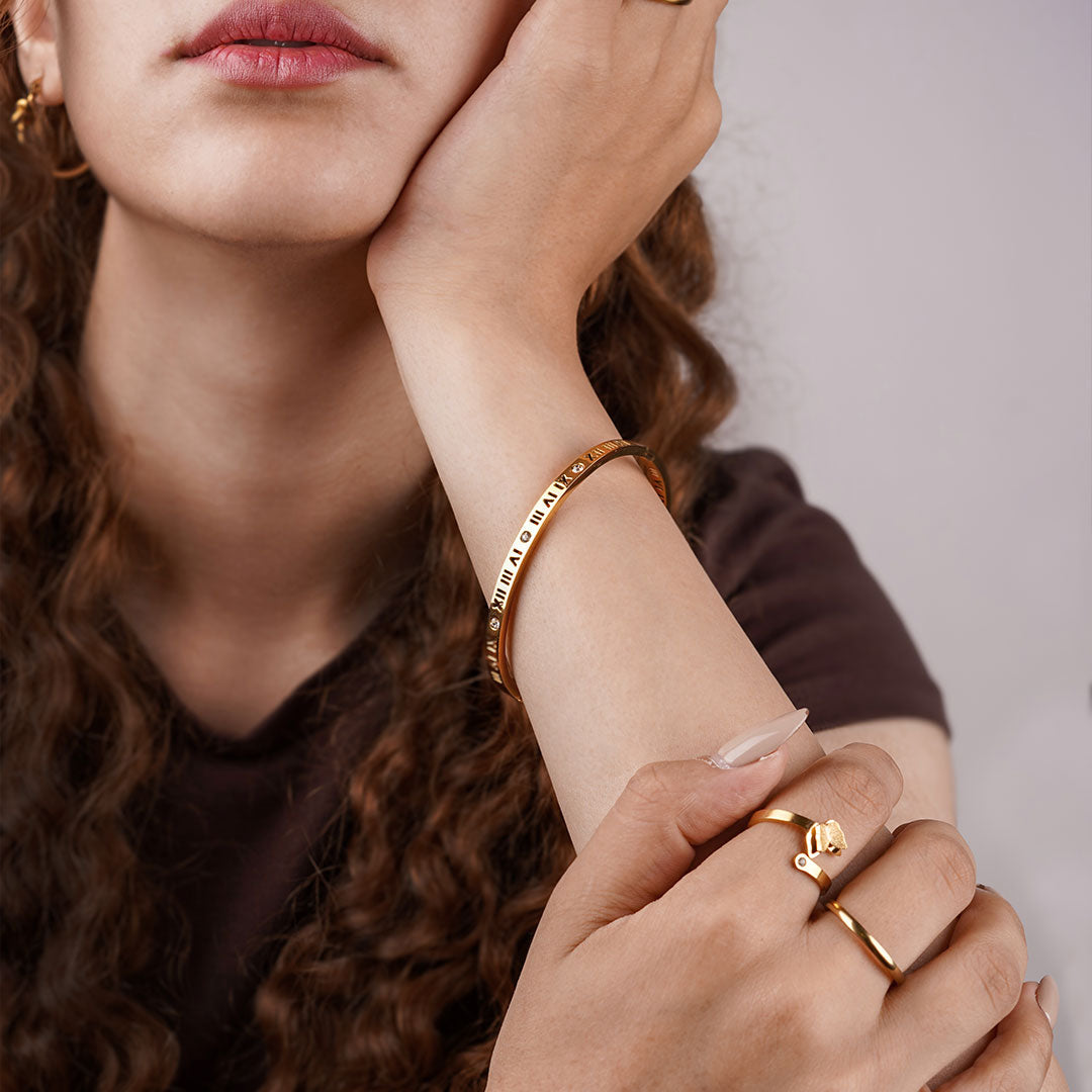 A person's arm is visible from the elbow down, wearing a polished gold bangle adorned with Roman numerals and a small, sparkling gemstone. The person is also wearing a gold ring with a butterfly charm and a simple gold band on their fingers.