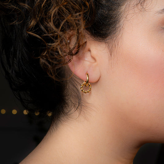 Close-up of a person's ear wearing a small, gold hoop earring. The hoop is smooth, and a smaller, textured, braided, or knot-like gold ring hangs from it.