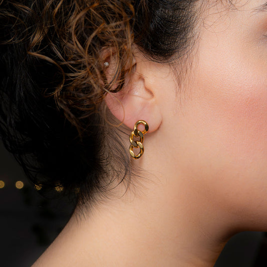 Close-up of a person's ear wearing a gold dangle earring, composed of three interconnected, shiny, oval-shaped chain links. The earring is called Delphine Shine Earrings.