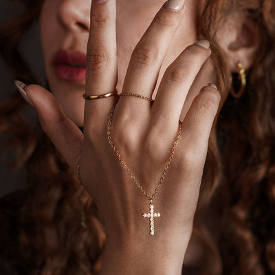 A close-up of a person's hand, adorned with a unique piece of jewelry. A thin gold chain connects a small, polished ring on the index finger to a delicate cross-shaped pendant, which is encrusted with small, clear gemstones.